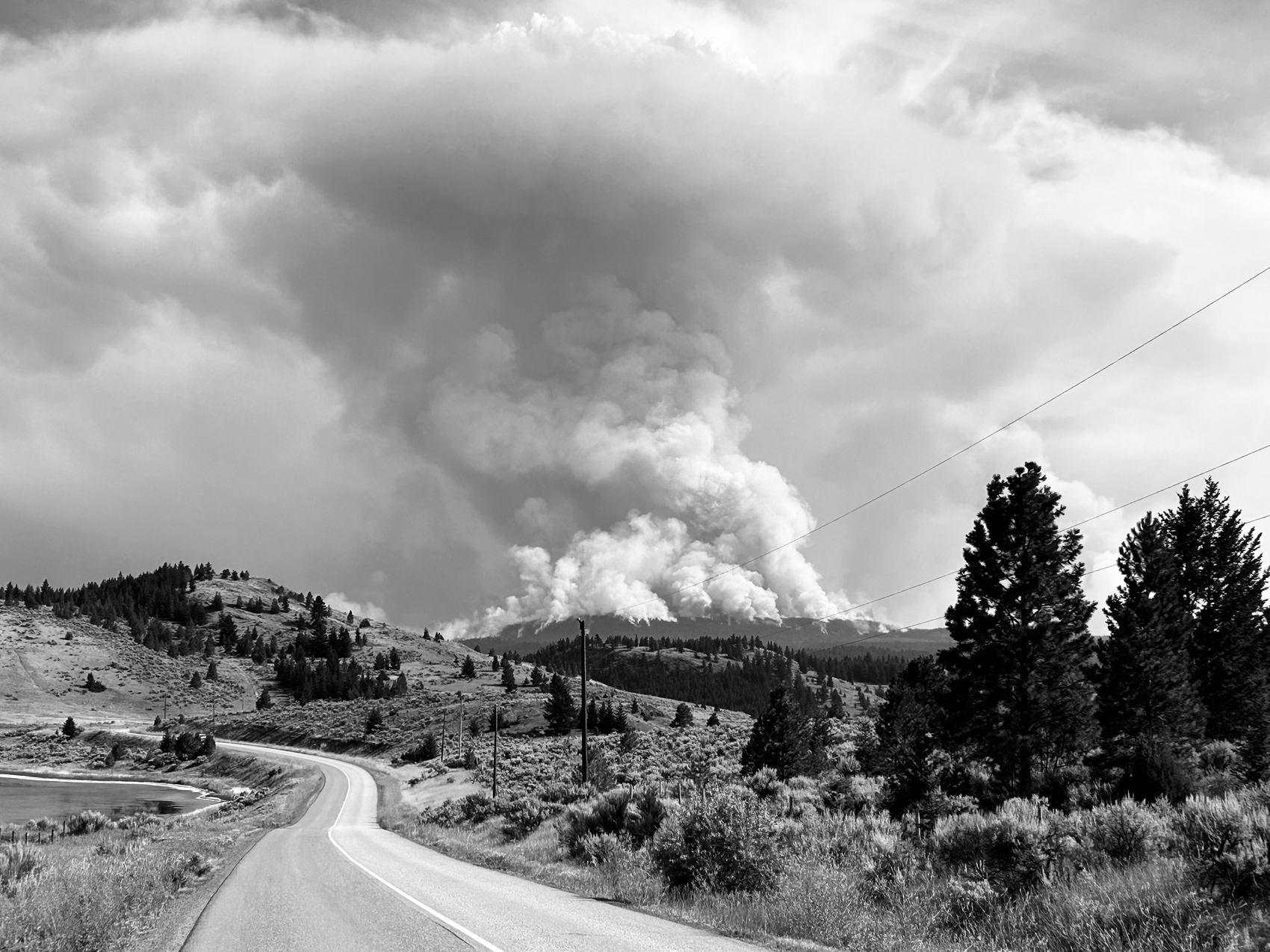 A road curving toward a wildfire smoke column rising over forested hills near Kamloops.